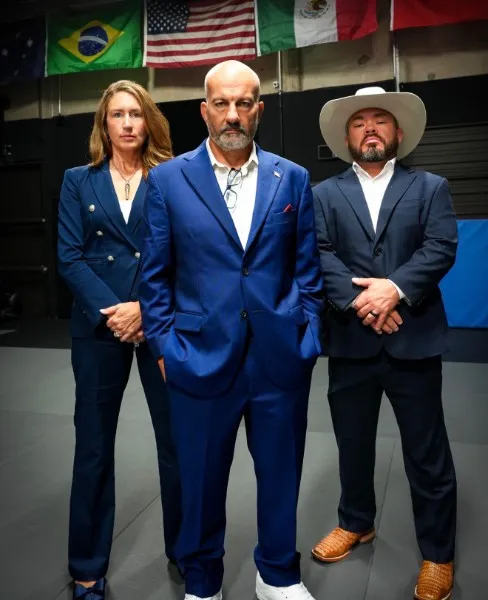Lindsay and Carlos Machado with Roger Perez, standing together in suits on the academy mats, under a row of national flags